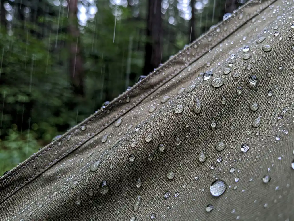 Waterdruppels die parelend van een geïmpregneerd canvas tent afrollen tijdens hevige regen, met een groen bos op de achtergrond.