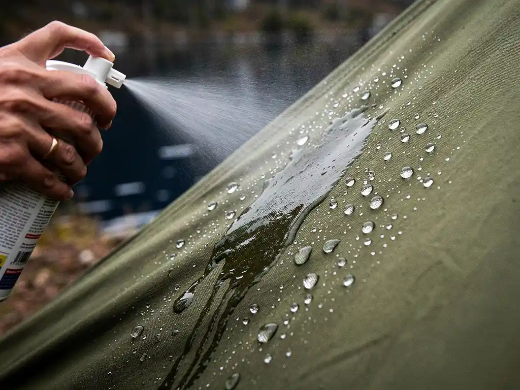 Waterparels die afpatten op behandeld canvas tentdoek na het aanbrengen van waterafstotende spray, mistig meercamp op de achtergrond.
